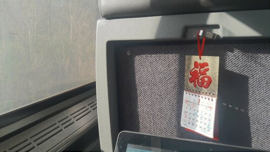 calendar with red 福 hanging on back seat. glimpse of computer on tray table out of view. trees outside are visible through gray window.