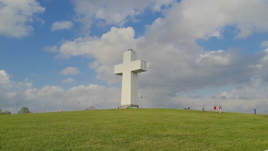 A large white cross on a green hill with blue cloudy sky and people nearby